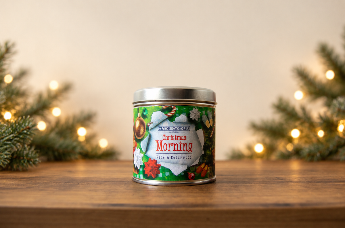 Clyde Candles Christmas Morning Pine & Cedarwood candle tin on a wooden table with evergreen branches and warm Christmas lights