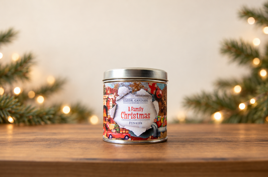 Clyde Candles A Family Christmas Fireside candle tin on a wooden table with evergreen branches and warm Christmas lights