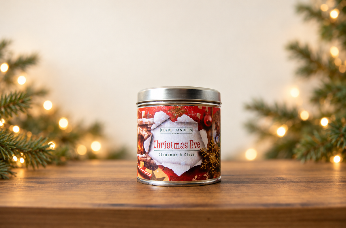 Clyde Candles Christmas Eve Cinnamon & Clove candle tin on a wooden table with evergreen branches and warm Christmas lights
