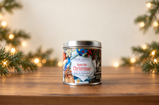 Clyde Candles Home for Christmas Apple & Cinnamon candle tin on a wooden table with evergreen branches and warm Christmas lights